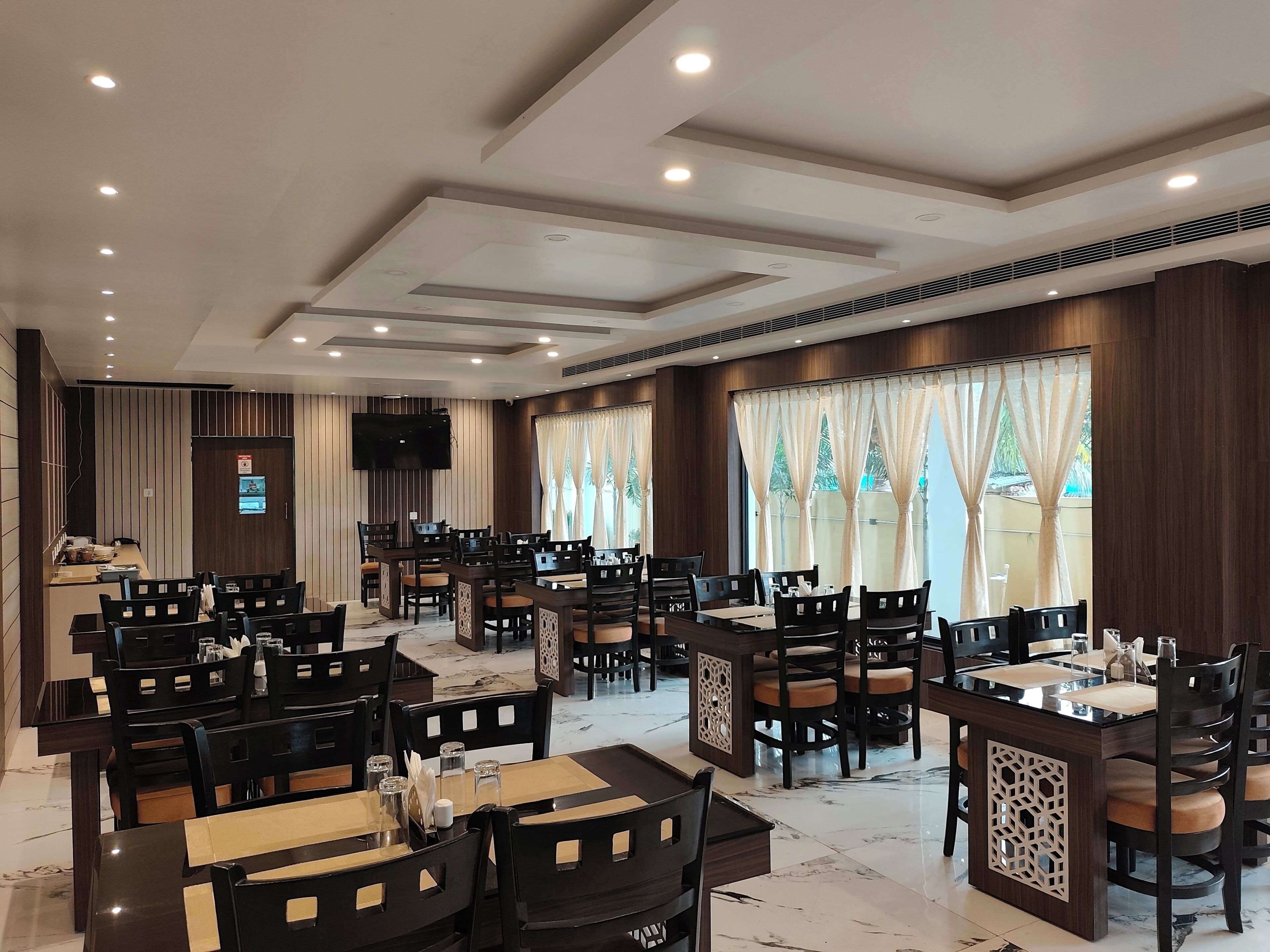 A hotel dining area with multiple tables and chairs, a flat-screen TV mounted on the wall, and large windows with curtains.
