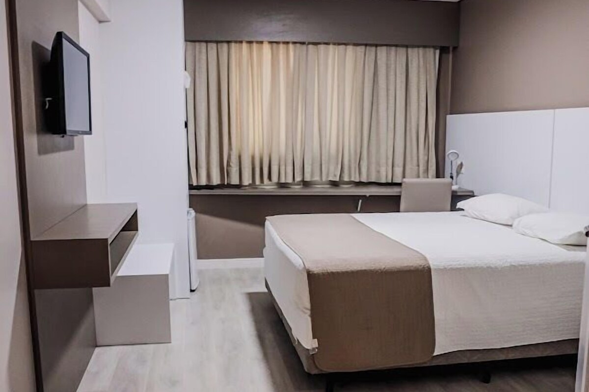 A modern hotel room with a bed, bedside table, TV, and a window with curtains.