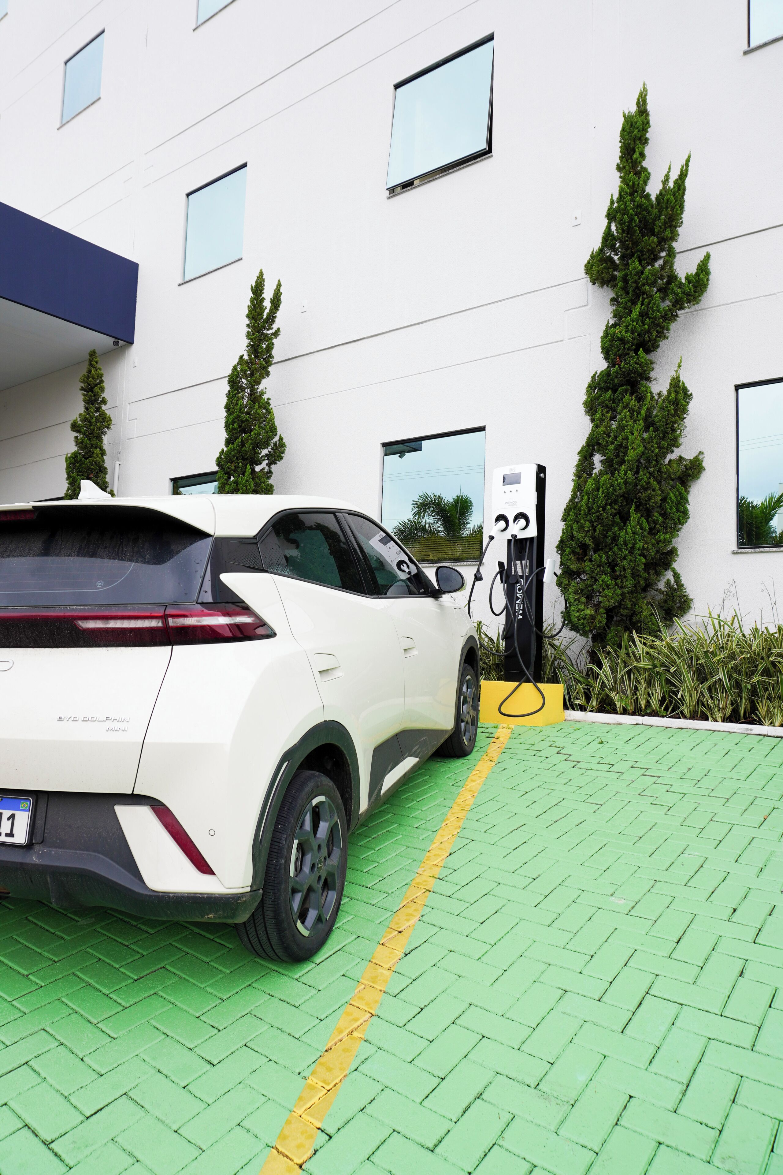 Un vehículo eléctrico blanco cargándose en una estación frente a un edificio con múltiples ventanas y jardines.