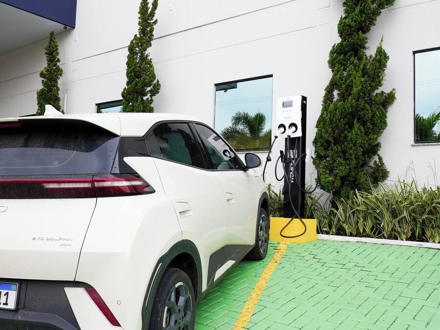 Un vehículo eléctrico blanco cargándose en una estación frente a un edificio con múltiples ventanas y jardines.