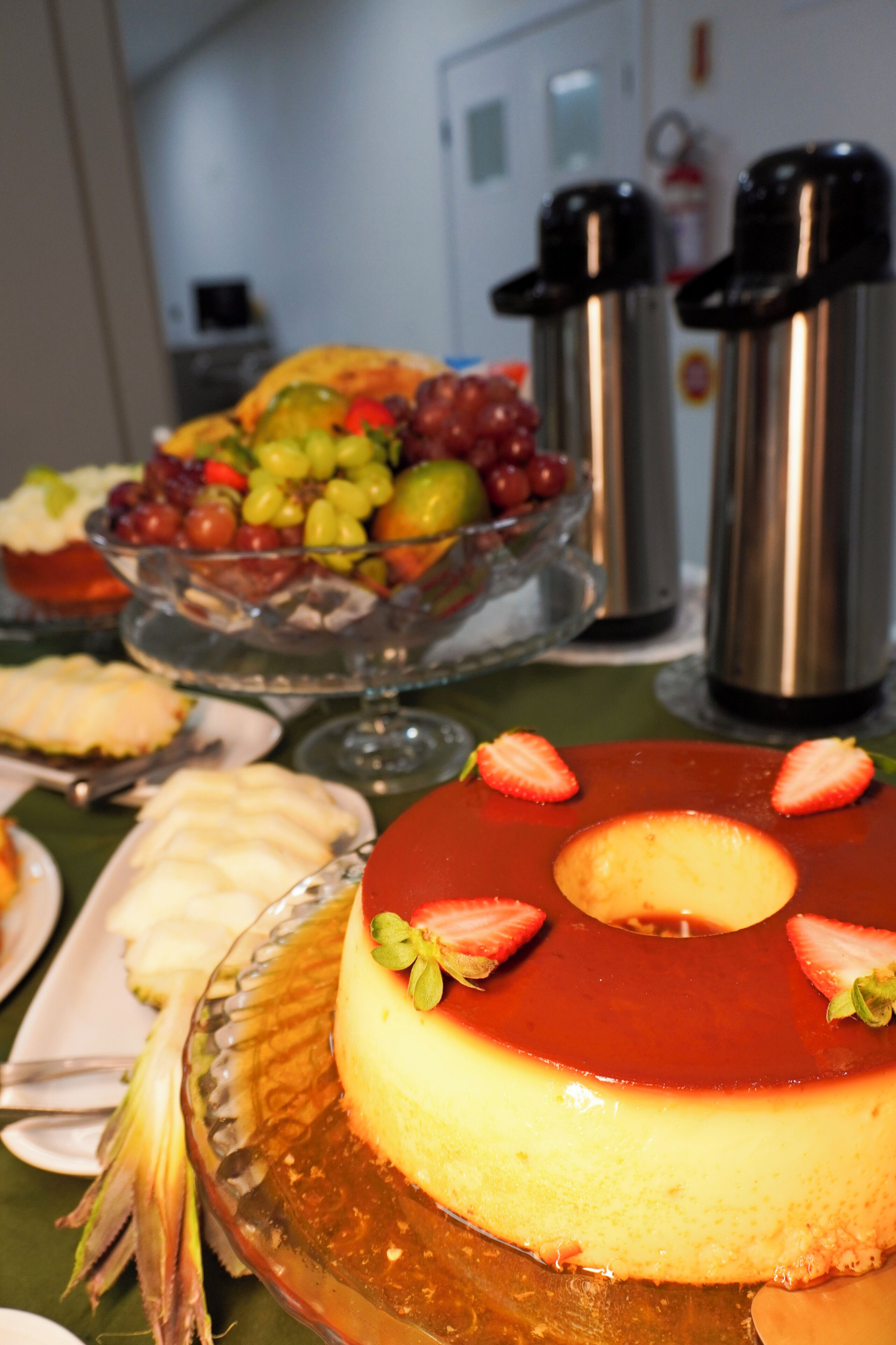 A dessert table with a flan, fruit bowl, and coffee makers.