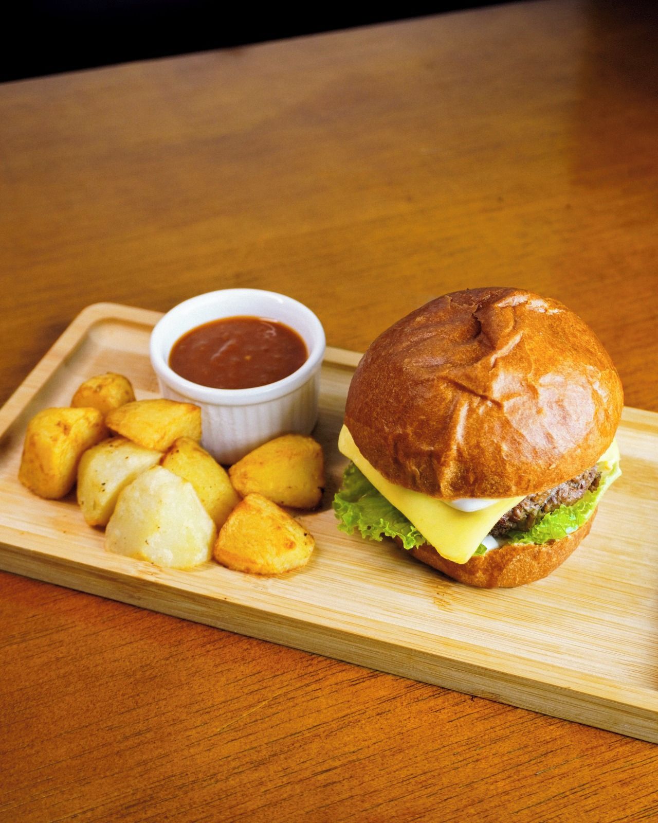 A meal consisting of a burger with lettuce and cheese, a side of golden potato wedges, and a small bowl of red sauce on a wooden tray.