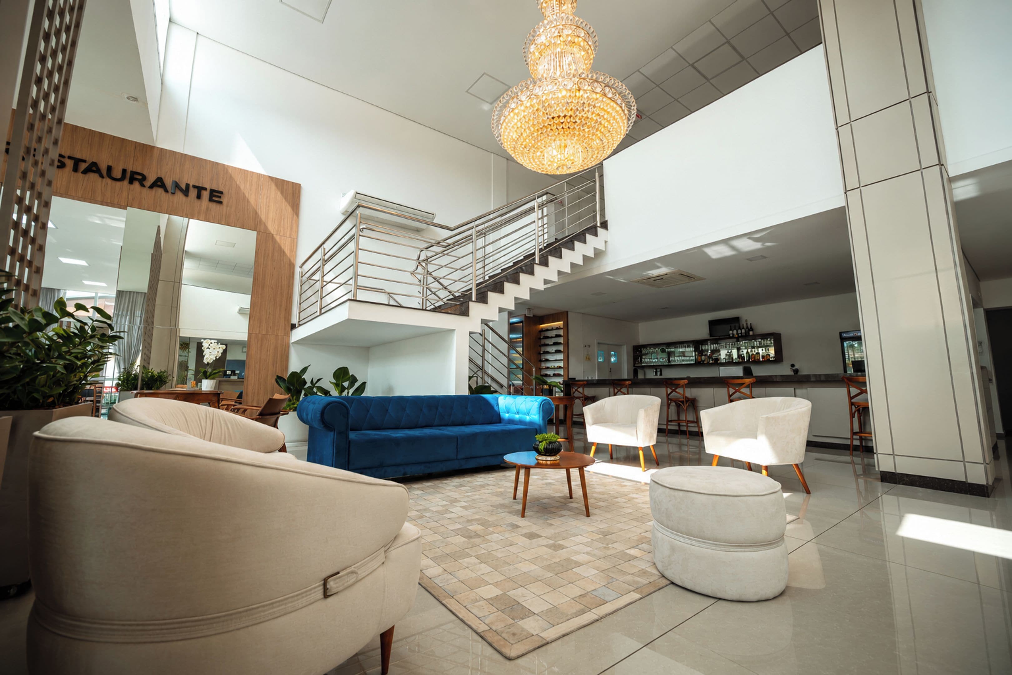 Ampla sala de estar com sofá azul, poltronas brancas e um lustre. Uma escada leva a um andar superior.