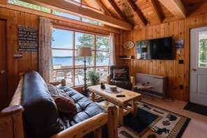 Ein gemütliches Blockhaus mit Holzvertäfelung, einem großen Fenster mit Seeblick, einer bequemen Couch, einem Fernseher und einem Kamin.