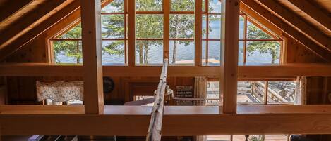 Une cabane avec une chambre à coucher située en mezzanine, de grandes fenêtres, une échelle en bois et une vue sur un quai et un lac.