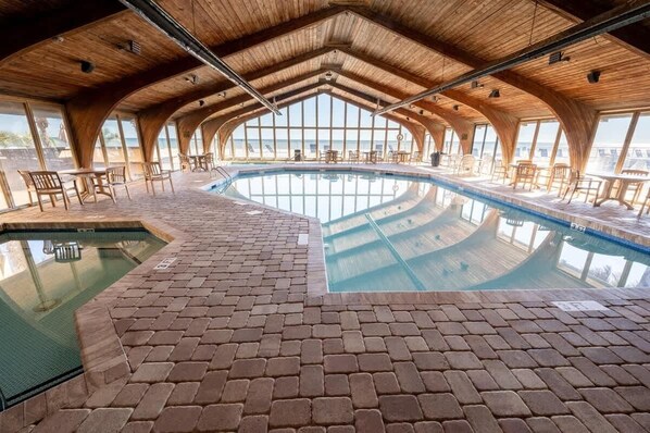Una piscina interior con suelo de ladrillo y techo de madera, rodeada de sillas y mesas.