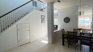 Uma sala de estar ampla com escada, mesa de jantar com cadeiras, relógio na parede e janela que oferece luz natural.