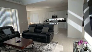 Uma sala de estar moderna com um sofá de couro preto, uma mesa de centro de madeira e um tapete estampado. Há uma área de cozinha com armários brancos e um micro-ondas.