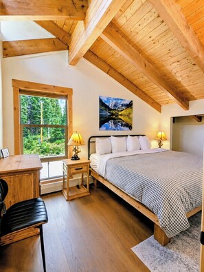 Een slaapkamer met een houten plafond, een bed met witte lakens, een schilderij van een berglandschap en een raam met uitzicht op groen.