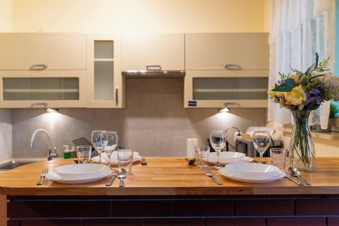A modern kitchen with a wooden countertop, two white plates, wine glasses, and a vase with flowers.