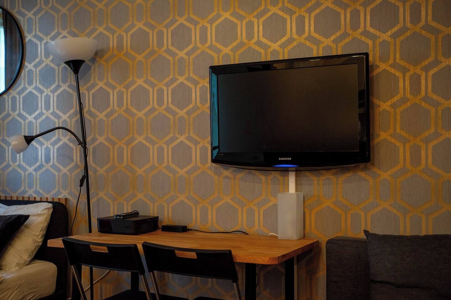 Una habitación con televisor de pared, escritorio con silla, lámpara y sofá.