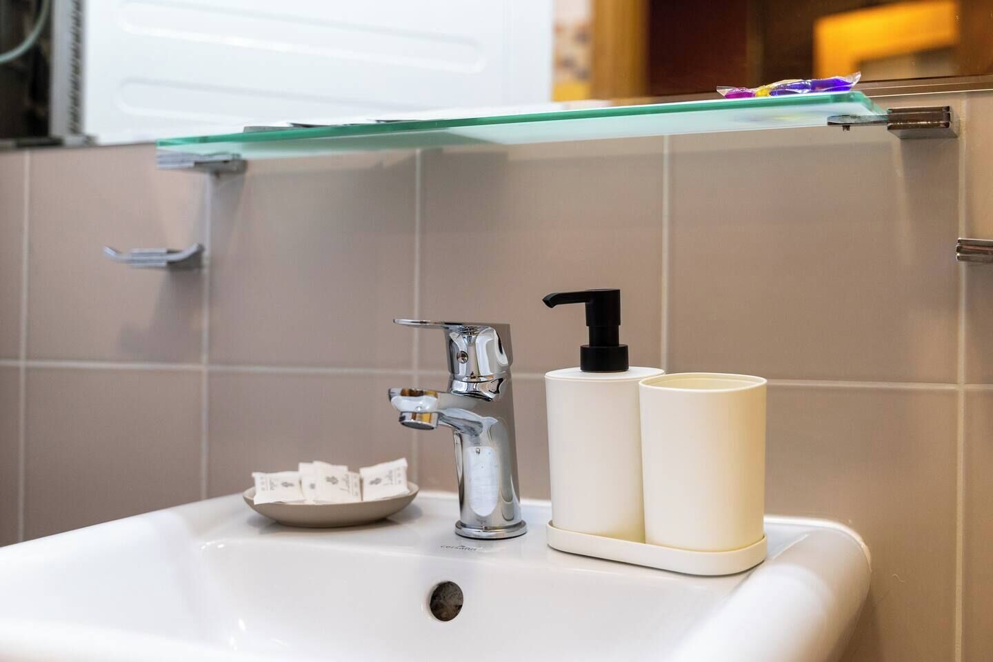 A bathroom sink with a chrome faucet, a soap dispenser, and a glass shelf above.