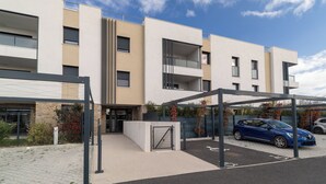 A modern multi-story building with a covered parking area and a blue car parked inside. - L’Hortus – Vue montagnes - Balcon (Saint-Mathieu-de-Tréviers)