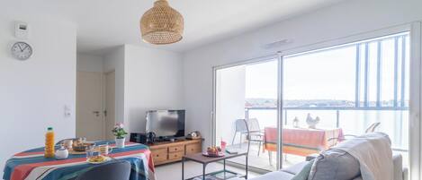 A modern living room with a sofa, a dining table with a colorful tablecloth, a television, and a balcony with a view.
