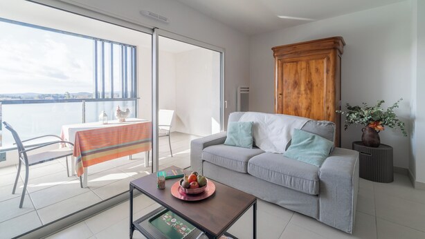 A modern living room with a grey sofa, a wooden cabinet, a dining table with a striped tablecloth, and a balcony with a view of the city. - L’Hortus – Vue montagnes - Balcon (Saint-Mathieu-de-Tréviers)