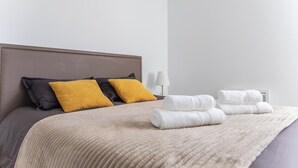 A neatly made bed with a beige blanket, two pillows in black and yellow, and two folded white towels. - L’Hortus – Vue montagnes - Balcon (Saint-Mathieu-de-Tréviers)