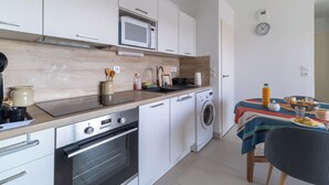 A compact kitchen with white cabinets, a microwave, a sink, a stove, and a washing machine. There's a table with a striped tablecloth and a chair. - L’Hortus – Vue montagnes - Balcon (Saint-Mathieu-de-Tréviers)