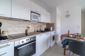 A compact kitchen with white cabinets, a microwave, a sink, a stove, and a washing machine. There's a table with a striped tablecloth and a chair. - L’Hortus – Vue montagnes - Balcon (Saint-Mathieu-de-Tréviers)
