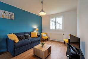 Un salon avec un canapé bleu, des coussins jaunes, une table basse en bois, un fauteuil jaune, une télévision et une fenêtre donnant sur des immeubles.