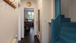 Un couloir avec un escalier bleu, une horloge et une porte donnant sur un espace cuisine.