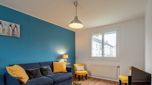 Un salon avec un mur bleu, un canapé orné de coussins jaunes, un fauteuil jaune, une lampe de table et une fenêtre donnant sur des immeubles.