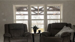 Een kamer met twee fauteuils, een kleine tafel met een plant en een kopje, en een groot raam met uitzicht op bomen.