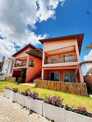Un edificio residencial de dos pisos con un balcón, cerca viva de madera y un jardín con flores rosas.