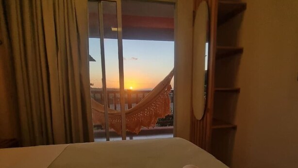 A hotel room with a bed, a view of the sunset through a window, and a hammock outside. - Beachfront house with sunset views (São Miguel do Gostoso)