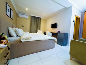 A hotel room with a bed, a desk, a chair, a TV, and an air conditioning unit.