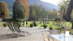 A wooden deck with chairs and a table, a playground, and a mountainous landscape in the background. - Seeidylle Winkler vacation apartment directly on Lake Ossiach with its own bathing beach (Bodensdorf)
