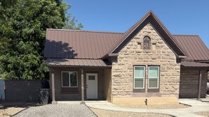 A two-story stone building with a brown metal roof, a front porch, and a gravel driveway.
