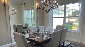 Un comedor con una mesa de madera dispuesta para seis personas, rodeada por seis sillas. Un candelabro cuelga sobre la mesa, y hay ventanas amplias que ofrecen vistas al exterior.