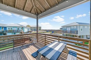 Une terrasse en bois avec une table et des bancs, donnant sur une plage avec des bâtiments en arrière-plan.