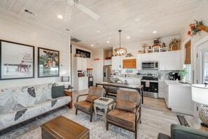 Un salon spacieux avec un canapé, deux fauteuils et une table basse. On aperçoit, au fond, une cuisine avec des meubles blancs et des appareils électroménagers.