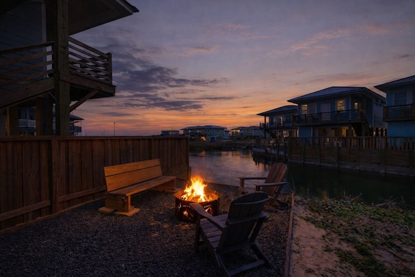 Una terraza de madera con una fogata, asientos y vistas a casas al otro lado del agua al anochecer.