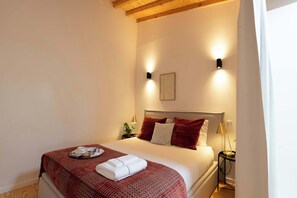 Ein ordentlich gemachtes Bett mit roter Decke und weißen Kissen, ein Nachttisch mit einer Lampe und eine Wandleuchte.