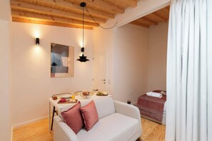 Ein gemütliches Zimmer mit Holzbalkendecke, einem weißen Sofa mit rosafarbenem Kissen, einem Esstisch mit Frühstücksutensilien und einem Bett mit rot gemustertem Überwurf.