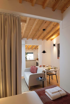 Ein gemütliches Zimmer mit einem Bett, einem kleinen Tisch mit einer Tasse, einem Sessel und einer Essecke mit Tisch und Stühlen.