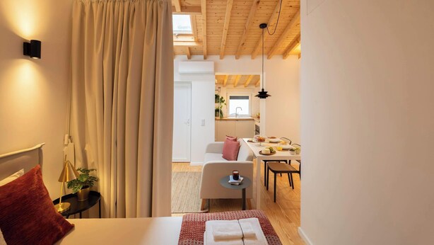 A modern hotel room with a bed, a bedside table with a lamp, a window with curtains, and a view of the kitchen and dining area. - Elegant Studio with Terrace & AC by HostWise (Porto)