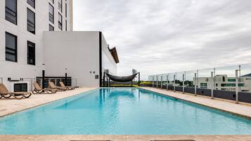 Una piscina en la azotea con sillas de descanso, una hamaca y un edificio moderno al fondo.