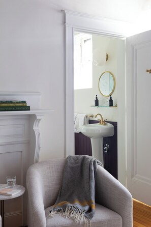 A room with a white fireplace, a round mirror above a sink, and a chair with a blanket. - West End Collection at 49 Neal (Portland)
