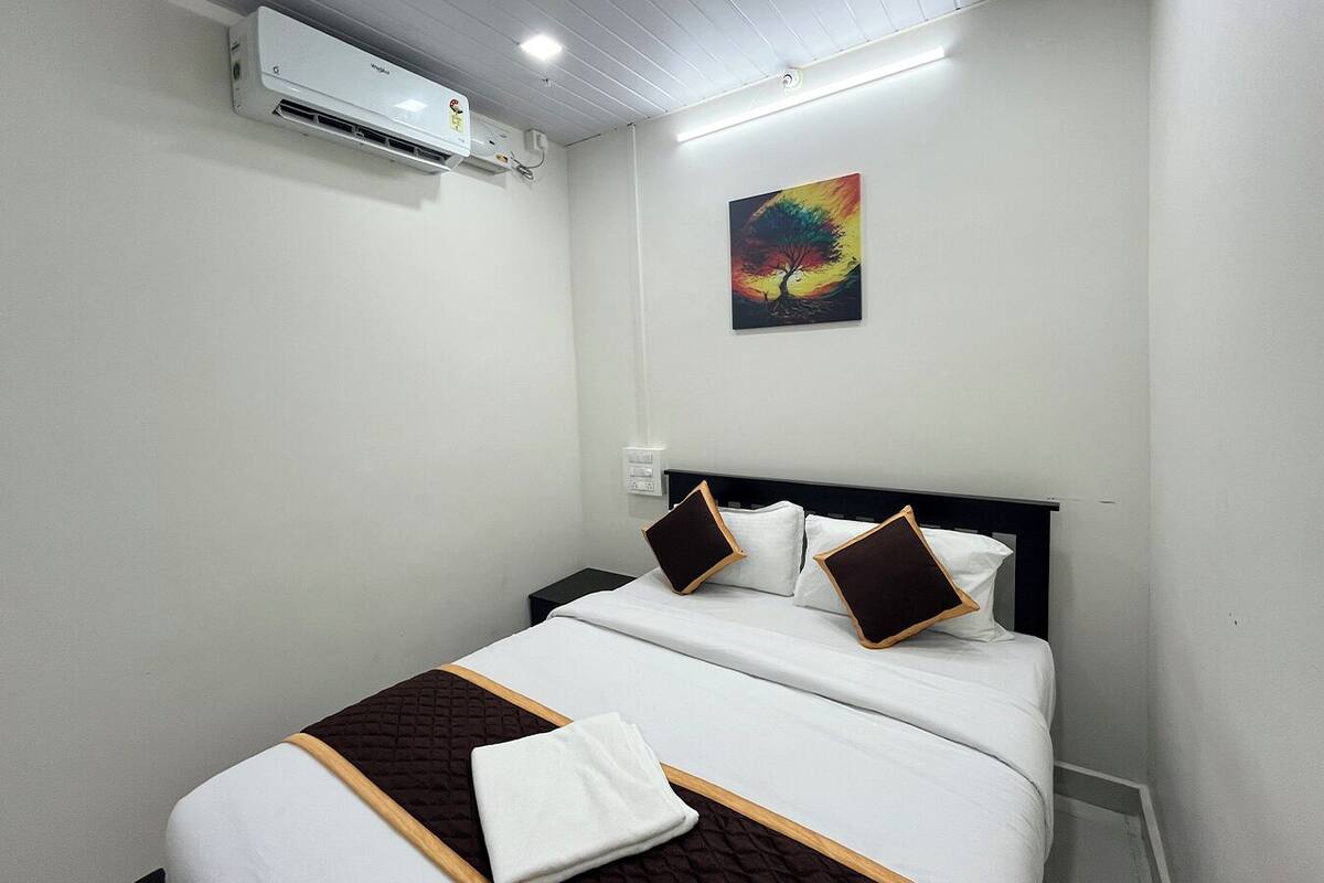 A hotel room with a bed, white walls, a painting, and an air conditioning unit.