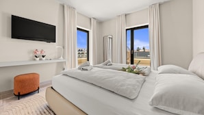 A modern hotel room with a large bed, a flat-screen TV, a small orange ottoman, and a view of the city through large windows. - Designer Beach Villa Limes - Pool, Spa & Games (Funtana)