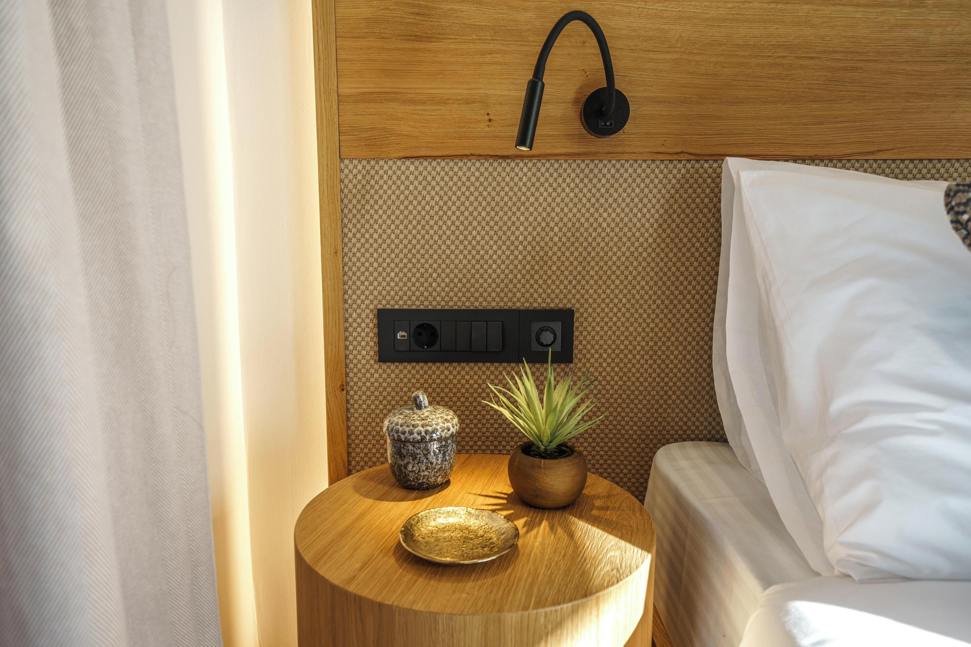 A neatly made bed with white sheets, a small wooden side table with a potted plant and a decorative bowl, and a wall-mounted light fixture.