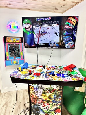 Un espace de jeu équipé d’un téléviseur, de jeux d’arcade et d’une table sur laquelle sont disposés divers accessoires de jeu.