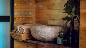 A wooden cabin interior with a stone basin, a wicker basket with towels, and a bottle of shower gel. - Ecolodge Cat Ba (Hai Phong)