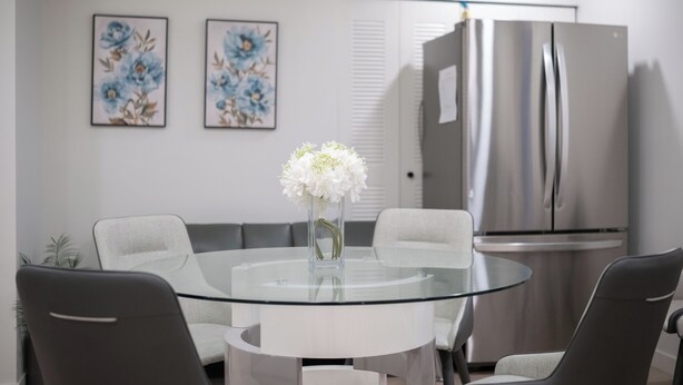 Una cocina moderna con un refrigerador de acero inoxidable, una mesa de vidrio con cuatro sillas y dos cuadros decorativos con motivos florales.