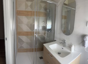 Un baño moderno con cabina de ducha de vidrio, un lavamanos blanco con secador de mano y un mueble de tocador de madera.