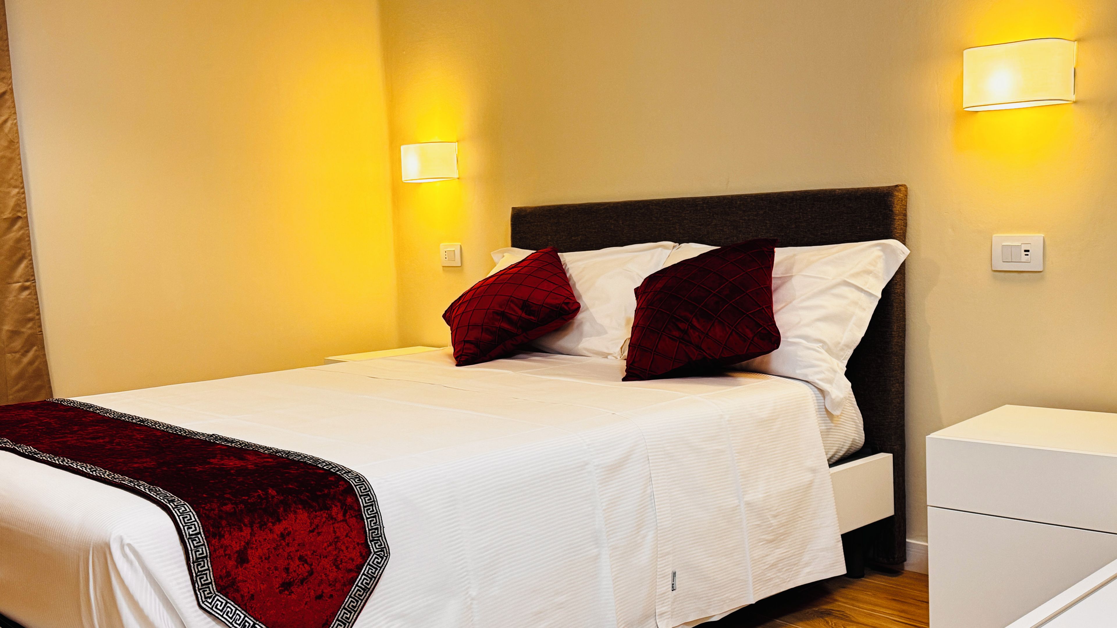 A neatly made bed with white linens and two red pillows, a bedside table, and a wall-mounted light fixture.