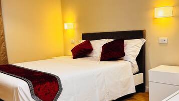 Una cama bien tendida con sábanas blancas y dos almohadas rojas, una mesita de noche y una lámpara fijada a la pared.